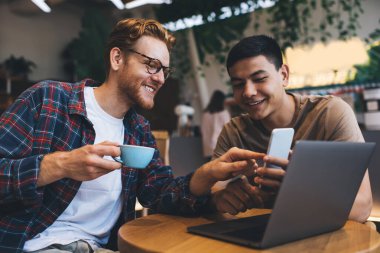 Kafkasyalı iş adamı Asyalı erkek arkadaşıyla izlerken akıllı telefonu işaret ediyor. İş birliği ve takım çalışması. Ofis masasında çalışan modern başarılı adamlar.