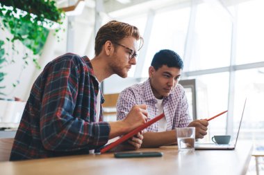 Asyalı iş adamı dizüstü bilgisayarında, ofiste yeni bilgiler yazan beyaz erkek arkadaşına bir şey gösteriyor. İş birliği ve takım çalışması. Masadaki genç ve başarılı adamlar