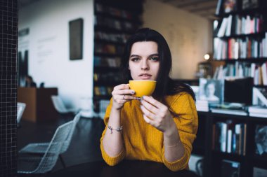 Kendine güvenen genç bir bayan modern çalışma alanında masa başında dinlenirken kameraya dalgın dalgın bakarak şık bir süveter ve bir fincan kahve giyiyor.