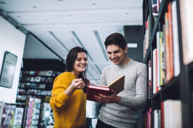 Modern kütüphanede birlikte yeni bir kitap seçerken sıcak süveter giyen, gülümseyen genç kadın ve erkeğin alçak açısı.