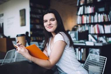 Ofiste masa başında oturmuş kameraya bakarken kitap okuyan ve buluşmayı bekleyen iyimser kadın çalışanlar.