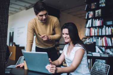 Beyaz tişörtlü pozitif genç bayan stajyer yaratıcı iş yerindeki konsantre erkek yöneticiye dizüstü bilgisayardan proje sonuçlarını gösteriyor.