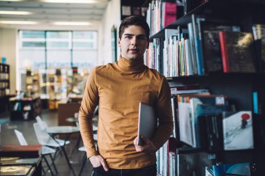 Çağdaş kütüphanede kitaplığın yanında duran ve elinde dizüstü bilgisayarla kameraya bakan düşünceli bir adam.