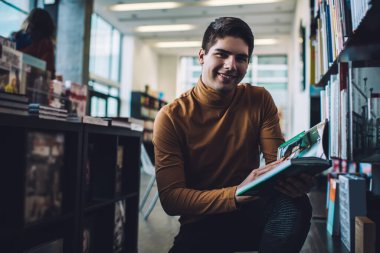 Günlük giysiler içinde gülümseyen genç adam kitap okurken kameraya bakıyor ve kütüphanede gülümsüyor.