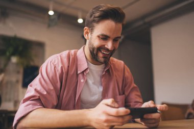 Neşeli hippi adam, arkadaşı beklerken Nintendo konsolu joystickiyle sanal 3D oynayarak eğleniyor. Gülümseyen erkek oyuncu, hafta sonu boş zamanlarında el bilgisayarı uygulaması üzerinden bir sonraki seviyeye geçiyor.