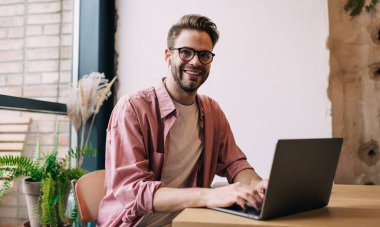 Optik gözlüklü neşeli bilişim uzmanı kafenin içinde uzaktan müzik dinlerken modern dizüstü bilgisayarda bluetooth 'a bağlı elektronik kulaklıklı mutlu bir erkek serbest çalışıyor.