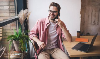 Uzaktan kumandalı alanda akıllı telefon sohbetinden keyif alan mutlu erkek serbest yazar. Gözlüklü neşeli hippi, netbook 'la masaya gülümsüyor. Aramak için cep telefonunu kullanıyor.