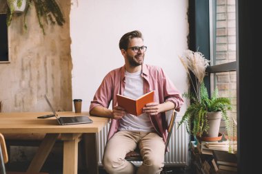 Dijital dizüstü bilgisayarı ve eğitim kitabı olan mutlu erkek öğrenci diğer tarafa bakıyor ve gülümsüyor, modern netbook ile Kafkasyalı serbest yazar ve birlikte çalışma ve sınavlara hazırlanıyor.