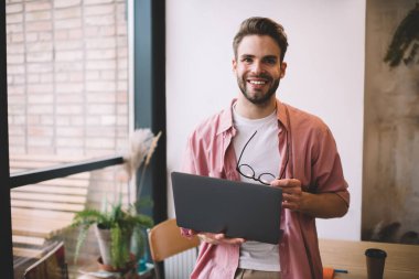 Diz üstü bilgisayarı ve kameradaki gözlüklerle iş arasında gülümseyen yarı uzunluktaki neşeli özgür erkek programcı dijital ağ kitabıyla eğleniyor.