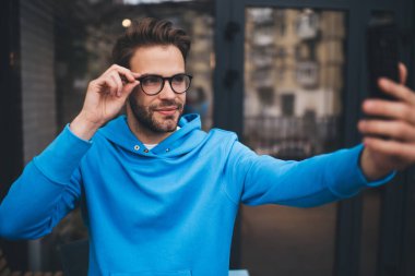 Klasik gözlüklü beyaz bir adam boş zamanlarında cep telefonu teknolojisi kullanarak selfie çekiyordu. Gündüz saatlerinde cep telefonunun ön kamerasına bakan yeniyetme bir adam.