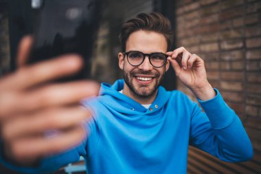 Gülümseyen hippi adam mobil teknolojiyle video bloğu çekiyor, komik milenyum erkek blogcusu akıllı telefon cihazıyla şehirde boş zamanlarında selfie çekerken 4G 'ye bağlanıyor.