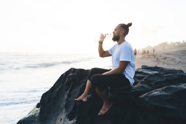 Sahildeki Rosky Stone 'da dinlenen erkek turistin yan görüntüsü ve boş zamanlarında vahşi sahili ziyaret etmek için su içmesi. Günlük kıyafetlerle yaz tatillerinin tadını çıkaran genç bir adam.