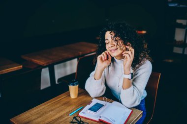 Kapalı gözlerle mutlu olan başarılı hippi kız kulaklıkla pozitif müzik listesi dinlerken ve hayal kurarken, kafeteryada müzik eşliğinde eğlenen kaygısız bayan öğrenci.