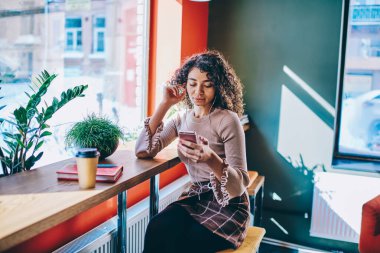 Büyüleyici hipster kızı akıllı telefondan video izliyor kulaklıkları ses oynatıcı olarak kullanıyor, ders kitabı olan bir blog yazarı cep telefonuyla takipçilerle sohbet ederken müzik dinliyor.