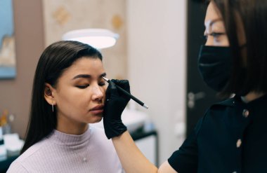 Güzellik uzmanı kuaförde Asyalı bir kızın iğnesiyle kaş mikropateni yapıyor. Kaşlar için yarı kalıcı makyaj. Kadın üstadın eldiven ve tıbbi maske taktığı kısmi görüntü. Güzellik konsepti
