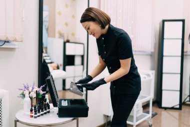 Kadın güzellik uzmanı kaş makyajı için iğne aletine ya da güzellik kutusundan mikroplara bakıyor. Eldiven takan gülümseyen Asyalı kadın üstat. Güzellik konsepti. Modern güzellik salonunun içi.