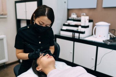 Güzellik uzmanı kuaförde Asyalı kız müşterinin iğnesiyle kaş mikro pateni yapıyor. Kaşlar için yarı kalıcı makyaj. Yetişkin kadın üstat eldiven ve tıbbi maske takıyor. Güzellik konsepti