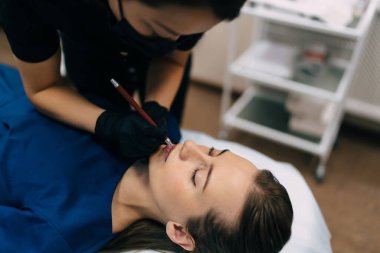 Güzellik uzmanı güzellik salonundaki beyaz kadın müşterinin dudaklarına şekil çiziyor. Kalıcı makyaj dudakları prosedürü. Eldiven ve tıbbi maske takan kadın üstadın kırpılmış görüntüsü. Güzellik konsepti