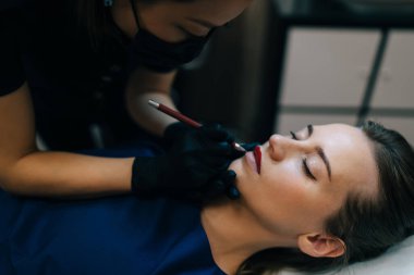 Güzellik salonundaki gözleri kapalı Avrupalı bir kızın dudaklarına hatlar çizen bir güzellik uzmanı imajı. Kalıcı makyaj dudakları prosedürü. Asyalı kadın üstat eldiven ve tıbbi maske takıyor. Güzellik konsepti