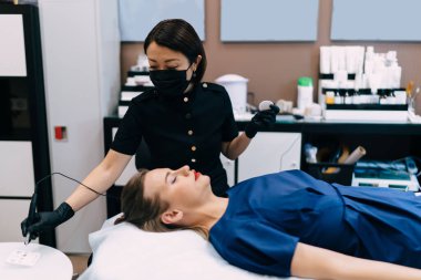 Güzellik uzmanı, kuaförde beyaz kadın müşteriye kalıcı makyaj yapmak için iğneyle ruj sürüyor. Eldiven ve tıbbi maske takan Asyalı kadın üstat. Güzellik konsepti