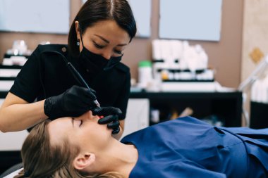 Güzellik uzmanı kuaförde gözü kapalı beyaz bir kız müşterinin iğnesiyle kalıcı makyaj dudak ameliyatı yapıyor. Asyalı kadın üstat eldiven ve tıbbi eldiven giyiyor. Güzellik konsepti