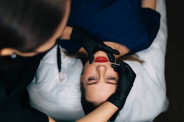 Güzellik uzmanının üst görüşü, güzellik salonundaki beyaz tenli kız müşterinin tahta çubuğuyla kalıcı makyaj dudakları prosedürü uyguluyor. Kadın üstadın eldiven ve tıbbi eldiven taktığı kısmi görüntü. Güzellik konsepti