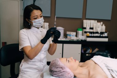 Güzellik uzmanı güzellik salonundaki kıza iğne yapmadan önce elinde botoks iğnesiyle şırıngaya bakıyor. Genç beyaz kadın müşteri tıbbi şapka takıyor. Güzellik konsepti. Kadın üstat eldiven ve tıbbi maske takıyor.