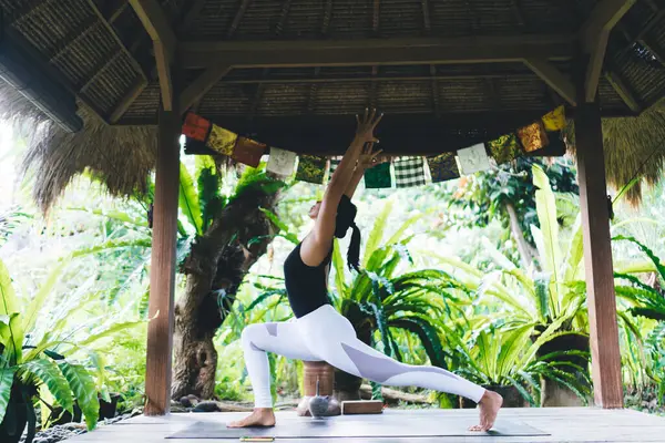 stock image Side view of girl practicing yoga in Warrior 1 pose. Concept of healthy lifestyle. Young athletic woman wearing sportswear and barefoot on fitness mat on wooden terrace. Bali island. Sunny day