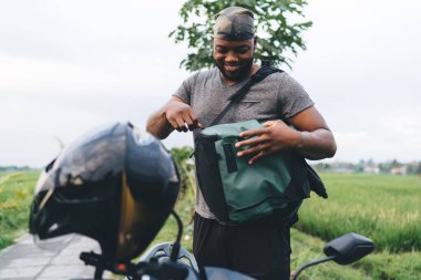 Bali 'de sırt çantasında bir şey ararken scooter' ın yanında gülümseyen neşeli Afrikalı Amerikalı turist. Bandanalı mutlu koyu tenli hippi Endonezya 'da yaz tatilinin tadını çıkarıyor.