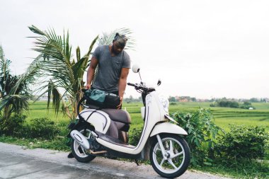 Eski model scooter 'ın yanında duran genç erkek turist tropikal palmiye ve patikanın yanına park etmiş, Endonezya' da seyahat sırasında eski mobiletiyle seyahat eden Afrikalı Amerikalı hippi.