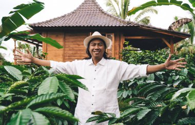 Endonezya 'da kahve çalısı ile tarımsal çevrede kameraya gülümseyen sıradan şapkalı mutlu Balili erkek, kafein bitkileriyle tarlalarda poz veren neşeli işadamı.
