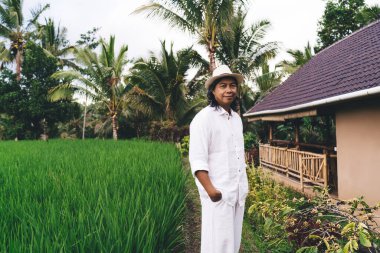 Tayland 'da gündüz vakti kameraya bakan sıradan şapkalı Balili çiftçinin yarım boy portresi, kırsal kesimde pirinç tarlalarıyla poz veren beyaz giysili yetişkin bir adam - doğal ekoloji.