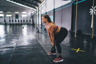 Spor salonunda antrenman yaparken kettlebell eşofmanlı, kaslı bayan vücut geliştirici, güçlü beyaz tenli sporcu, kavşak egzersizi sırasında spor halteri ekipmanı kullanıyor.