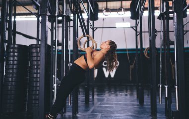 Spor eşofmanlı beyaz bir sporcu spor yüzükleriyle barfiks çekiyor. Kadın dayanıklılığı ve vücut geliştirme motivasyonu kavramı. Kovalamaca enerjisi olan kaslı kadın eğitiminin yan görünüşü.