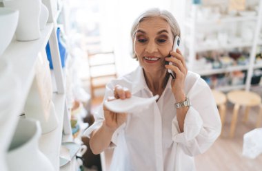 50 yaşında gülümseyen beyaz kadın girişimci akıllı telefondan konuşuyor ve seramik ürünlere bakıyor. Küçük işletmeler ve girişimcilik. Modern başarılı kadın. Raflarda çömlek olan ev sanatları stüdyosu