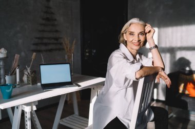 Sırıtkan Kafkas kadın girişimci sandalyeye oturmuş kameraya bakıyor. Küçük işletme ve girişimcilik kavramı. Karanlık sanat stüdyosunda güneşli ışıklandırması olan modern başarılı bir kadın.