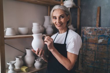 50 yaşındaki kadın heykeltıraş elinde seramik kap tutarak kameraya bakıyor. Küçük işletme ve girişimcilik kavramı. Ev hobisi, eğlence ve boş zaman fikri. Sanat stüdyosundaki kadın.