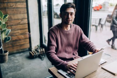 Kafe içinde modern dizüstü bilgisayar ve dijital cep telefonu teknolojisi kullanan Kafkasyalı hippi bir adamın portresi netbook ve akıllı telefonuyla kameraya bakan yetenekli bir erkek programcı.