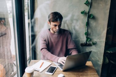 Milenyum erkek bilişim uzmanı elektronik kulaklıklarla laptop bilgisayarından indirme için video içeriğini araştırıyor, bluetooth kulaklıklarıyla beyaz blogcu web yayınlarını tarıyor.