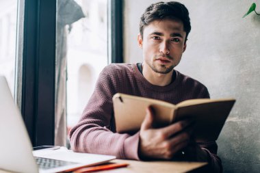 Egitim defteriyle birlikte çalisma alaninda ögrenme zamaninda kameraya bakan erkek ögrenci portresi, ders kitabi ve dizüstü bilgisayari olan beyaz tenli adam içerik plani olusturuyor.