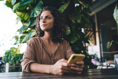 Kafe terasında dinlenen modern cep telefonu cihazıyla tefekkür eden kadın blogcu yayınlamak için web içeriğini düşünüyor, kıvırcık saçlı, dijital cep telefonu olan düşünceli hippi kız.