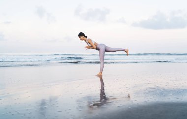 Mükemmel vücut yapısı olan beyaz kadın, sahil şeridinde ısınma sürecinde kaslarını geriyor. Çekici kadın, Asya yogasında meditasyon yaparken ve nefes alırken sağlıklı bir yaşam tarzına erişiyor.
