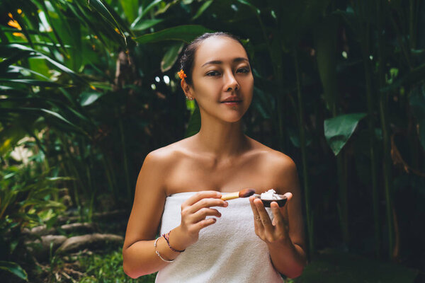Портрет привлекательной корейской женщины с кистью для макияжа и органическим кремом для очищения кожи и увлажнения на природе, очаровательной женщины, смотрящей в камеру, проводящей дневной спа для освежения тела