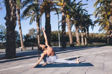 Yoga yaparken denge ve ahenk hisseden esnek bir kadın sokakta palmiye ağaçlarıyla paspas egzersizi yaparken asana pozu veriyor, aktif dişi yogi zindeliğini ve ruh sağlığını koruyor.