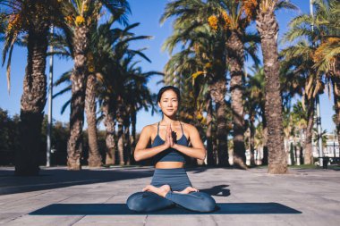 Aktif kıyafetli Asyalı kadın Lotus pozisyonunda oturuyor. Meditasyonu kolaylaştırmak için ellerini namaste pozisyonunda tutuyor. Yoga inziva sırasında palmiye ağaçlarıyla uyum içinde olan sakin bir kadın.