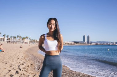 Sıska Asyalı kadın portresi. Gri tayt pantolon ve beyaz bluz. Hafta sonu okyanus kıyısında kameraya bakıyor.