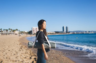 Spor giyim sektöründe spor yapan genç bir bayan antrenör deniz ufkuna bakarken hafta sonu kıyı şeridi antrenmanı için yer seçiyor. Çantalı çekici Asyalı kadın sağlıklı yaşam tarzını ve vücut pozitifliğini düşünüyor.