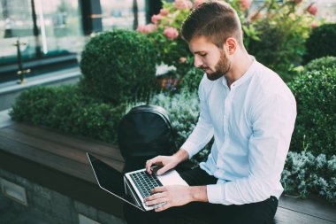 Yukarıdan, beyaz tişörtlü, siyah pantolonlu, sakallı, dizüstü bilgisayarının klavyesinde yazan, internette sörf yapan, bankta oturan genç bir adam.