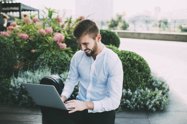 Beyaz gömlekli neşeli genç erkek, ahşap bankta oturmuş dizüstü bilgisayarını açıyor ve projede çalışırken ekrana bakıyor.