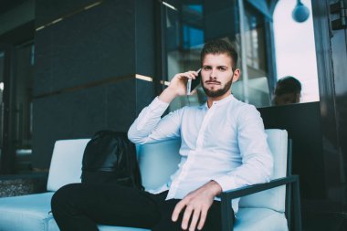 Beyaz gömlekli, siyah pantolonlu, koltukta oturmuş kameraya bakarken telefonla konuşan kendinden emin genç bir erkek.